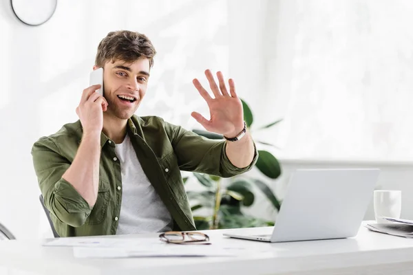 Bel homme d'affaires en chemise verte assis à la table avec ordinateur portable, parler sur smartphone et cinq Highing dans le bureau — Photo de stock