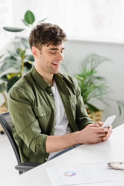 Bonito empresário em camisa verde sentado à mesa, sorrindo e segurando smartphone no escritório — Fotografia de Stock