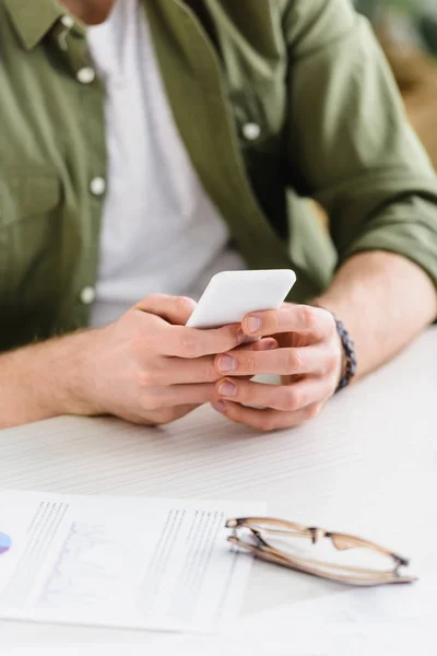 Cropped view of businessman in green shirt sitting at table and holding smartphone in office — Stock Photo