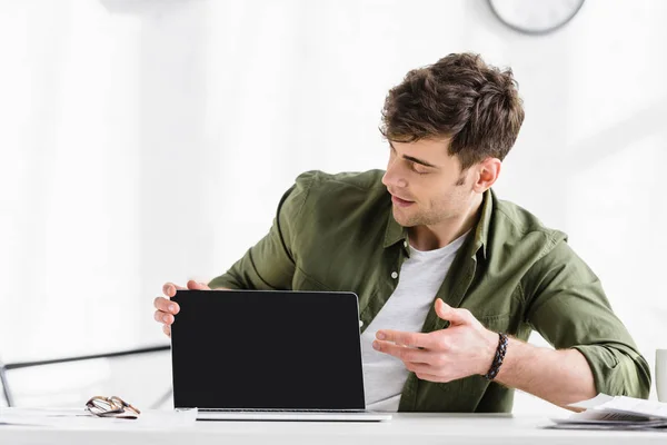Handsome businessman in green shirt sitting at table and pointing with hand at laptop with blank screen in office — Stock Photo