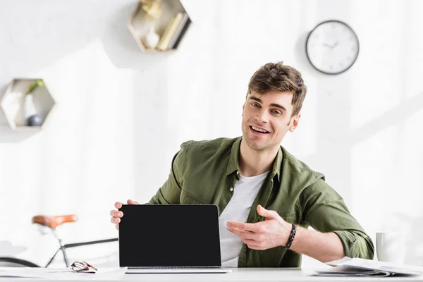 Hombre de negocios en camisa verde sentado a la mesa y apuntando con la mano a la computadora portátil con pantalla en blanco en la oficina - foto de stock