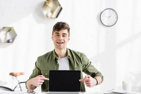 Bel homme d'affaires en chemise verte assis à table et pointant des doigts à l'ordinateur portable avec écran blanc dans le bureau — Photo de stock