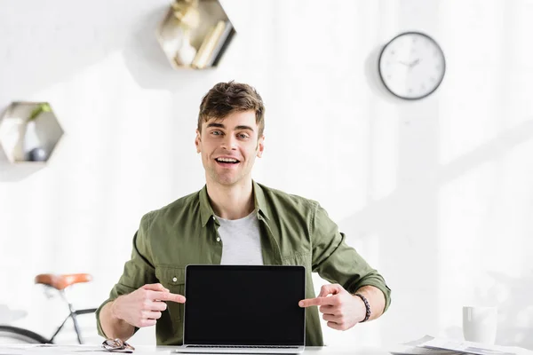 Hombre de negocios guapo en camisa verde sentado a la mesa y apuntando con los dedos a la computadora portátil con pantalla en blanco en la oficina - foto de stock