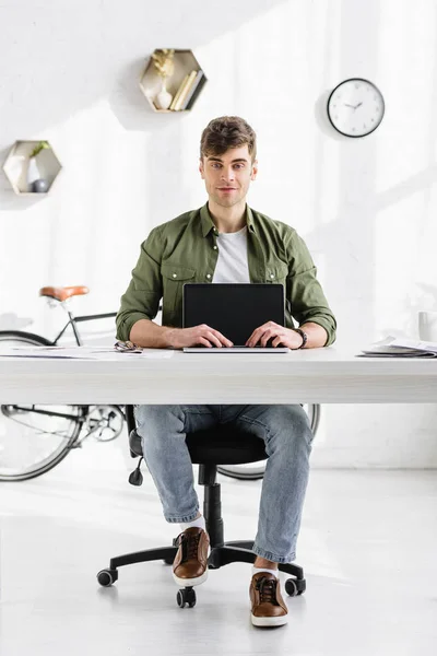 Handsome businessman in green shirt sitting at table and holding hands on laptop with blank screen in office — Stock Photo