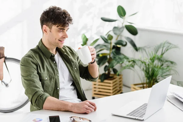 Bel homme d'affaires en chemise verte assis à table avec ordinateur portable et boire du café au bureau — Photo de stock