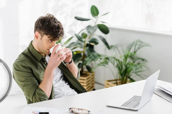 Hombre de negocios en camisa verde sentado en la mesa con el ordenador portátil y beber café en la oficina - foto de stock