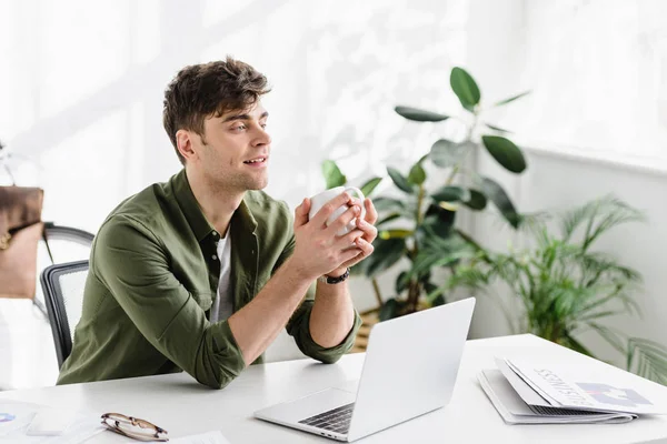 Handsome businessman in green shirt sitting, holding cup near table with laptop in office — Stock Photo