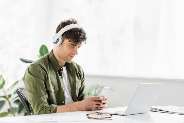 Schöner Geschäftsmann mit Kopfhörern sitzt am Tisch mit Laptop und hält Smartphone im Büro — Stockfoto