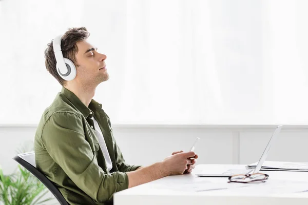 Schöner Geschäftsmann mit Kopfhörern sitzt am Tisch mit Laptop, hält Smartphone in der Hand und hört Musik im Büro — Stockfoto