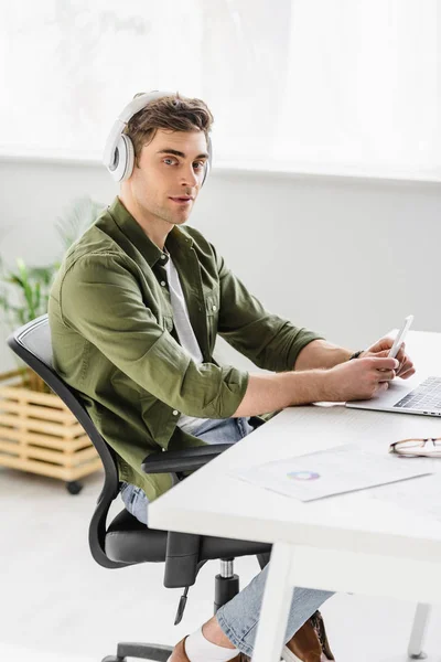 Hombre de negocios guapo en los auriculares sentados en la mesa con el ordenador portátil, sosteniendo el teléfono inteligente y escuchando música en la oficina - foto de stock