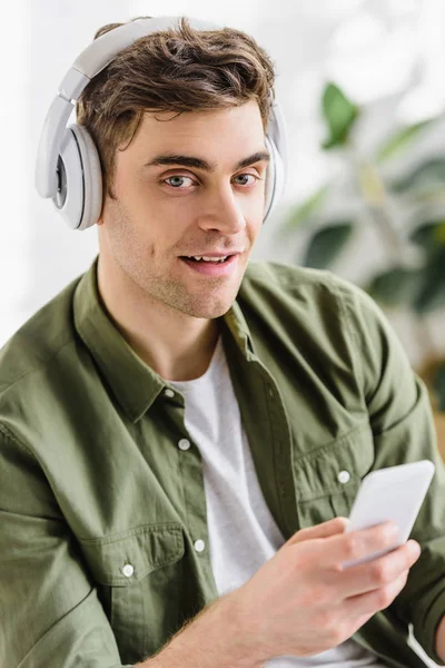 Homem de negócios bonito em camisa verde e fones de ouvido segurando smartphone no escritório — Fotografia de Stock