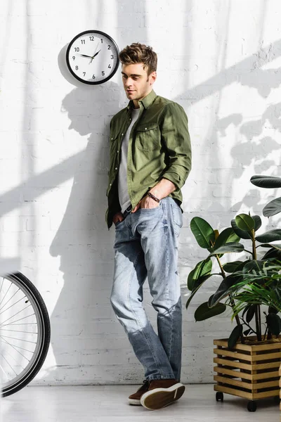 Bel homme en chemise verte et jeans debout près du mur de briques, vélo et plantes dans le bureau — Photo de stock