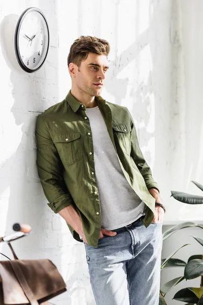 Hombre guapo en camisa verde y jeans de pie cerca de la pared de ladrillo, reloj y plantas en la oficina - foto de stock