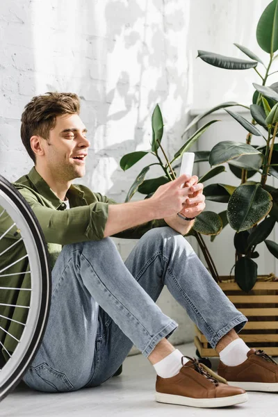 Hombre guapo en camisa verde y jeans con teléfono inteligente, sentado cerca de la pared de ladrillo, bicicleta y planta en la oficina - foto de stock