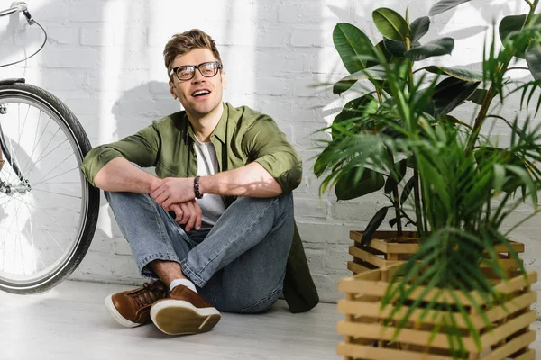 Hombre guapo en camisa verde, jeans y gafas sonriendo y sentado en el suelo cerca de la pared de ladrillo, bicicleta y plantas en la oficina - foto de stock