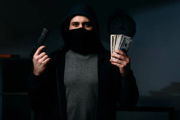 Thief in mask holding gun and dollar banknotes in dark room — Stock Photo