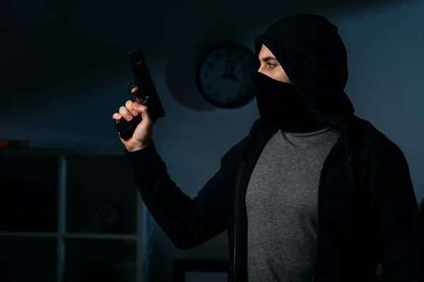 Maskierter Einbrecher hält Pistole im dunklen Raum und schaut weg — Stockfoto
