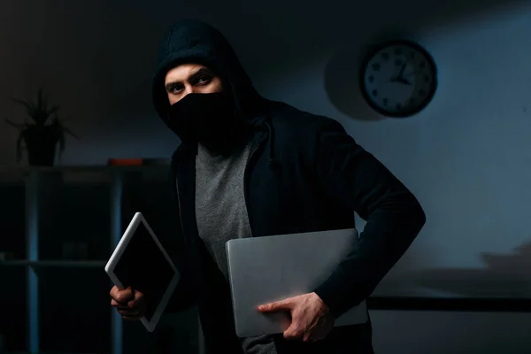 Thief in mask stealing laptop and digital tablet and looking away — Stock Photo