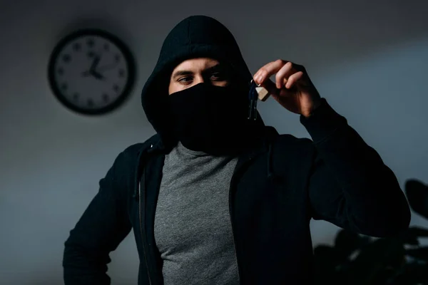 Confident thief in mask and hoodie standing in dark room and looking at keys — Stock Photo