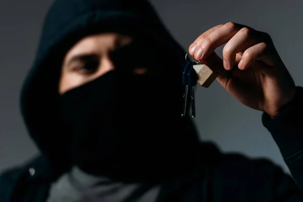 Thief in mask and black hoodie looking at keys on grey — Stock Photo