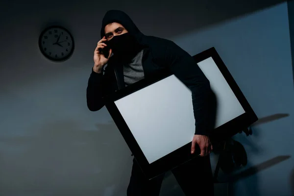 Thief in mask talking on smartphone while stealing flat-screen tv with blank screen — Stock Photo