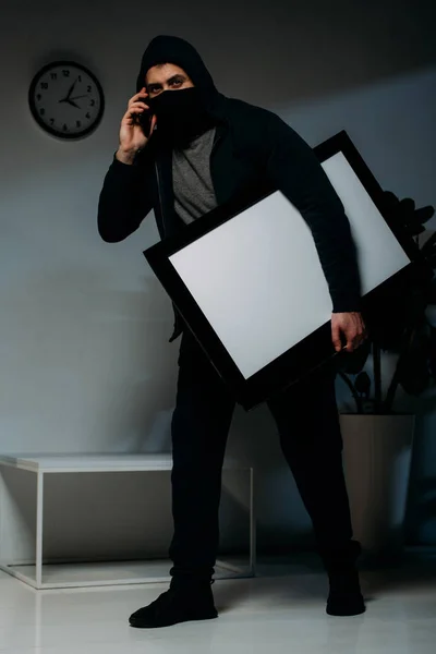 Ladrão mascarado falando no smartphone enquanto rouba tv de tela plana com tela em branco — Fotografia de Stock