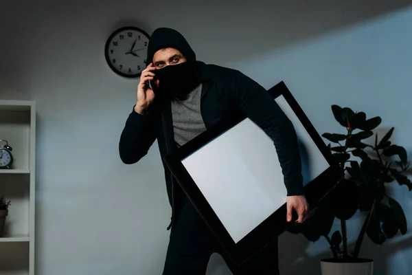 Ladrón en la máscara hablando en el teléfono inteligente mientras que el robo de televisión de pantalla plana con pantalla en blanco - foto de stock