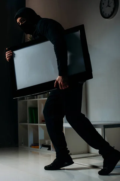 Cautious thief in mask stealing flat-screen tv with blank screen — Stock Photo