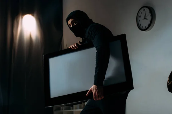 Robber in black mask stealing flat-screen tv with blank screen from apartment — Stock Photo