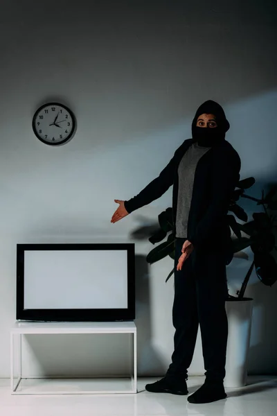 Ladrón sorprendido con máscara negra apuntando con las manos a la televisión de pantalla plana con pantalla en blanco - foto de stock