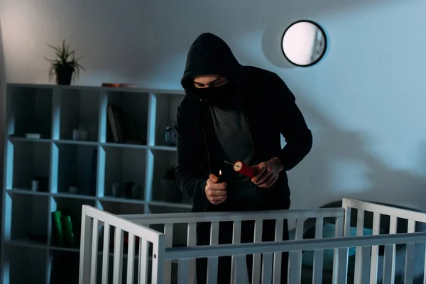 Criminal in mask igniting dynamite near crib in dark room — Stock Photo