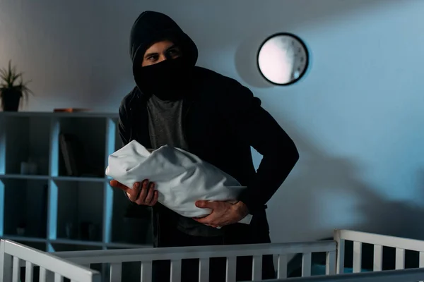 Похититель в черной маске держит младенца возле кроватки — стоковое фото