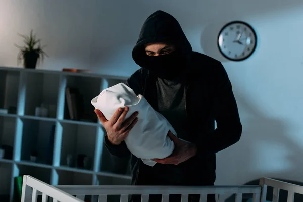 Kidnapper in mask and black hoodie standing near crib and holding infant child — Stock Photo