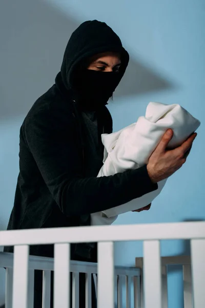 Kidnapper in mask standing beside crib and holding infant child — Stock Photo