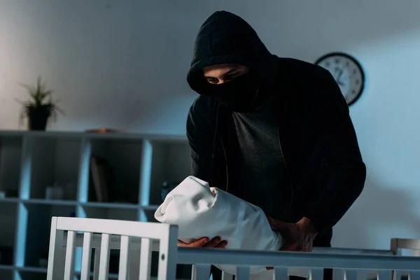 Secuestrador con máscara y sudadera con capucha de pie cerca de la cuna y sosteniendo al bebé - foto de stock