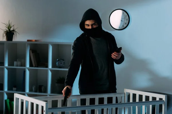 Преступник в маске держит смартфон и целится пистолетом в кроватку — стоковое фото