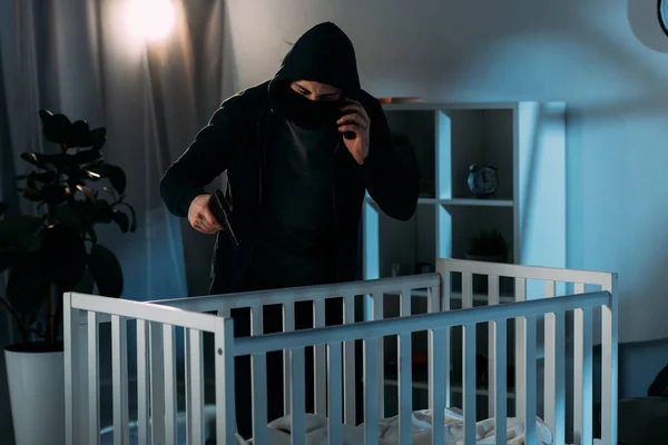 Criminal in mask talking on smartphone and aiming gun in crib — Stock Photo