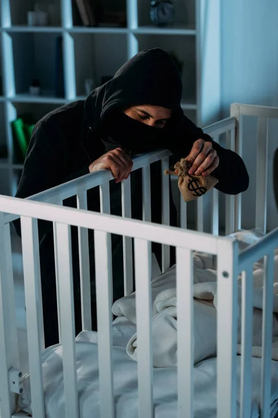 Secuestrador en máscara y sudadera con capucha negra sosteniendo bolsa de dinero y mirando en cuna - foto de stock