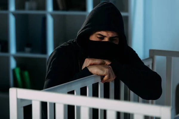 Sad kidnapper in mask and hoodie looking in crib — Stock Photo