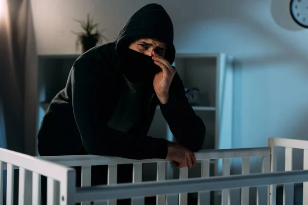 Crying kidnapper in mask standing near crib in dark room — Stock Photo