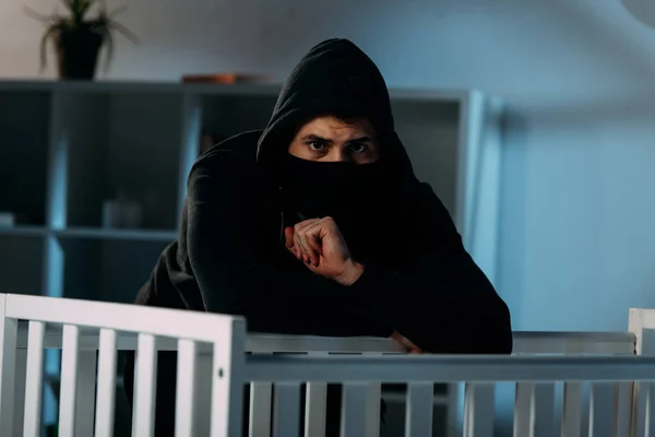 Worried kidnapper in black clothes and mask standing beside crib — Stock Photo