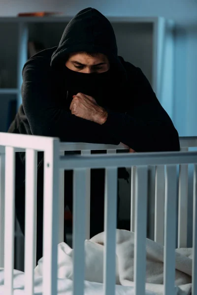 Pensive kidnapper in black clothes and mask looking in crib — Stock Photo