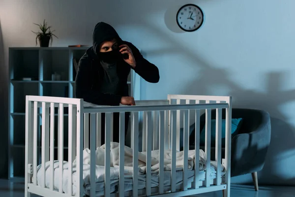 Kidnapper in black mask standing near crib and talking on smartphone — Stock Photo