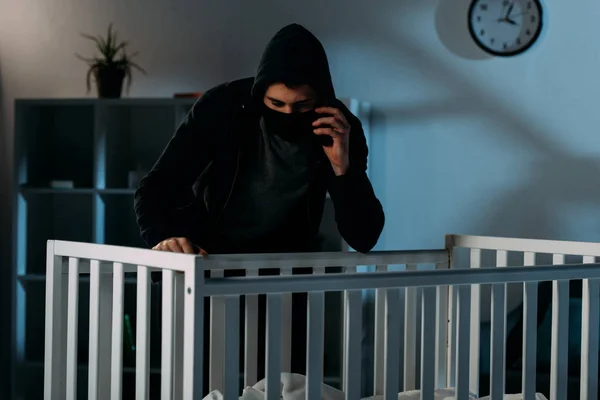 Kidnapper in mask talking on smartphone and looking in crib — Stock Photo