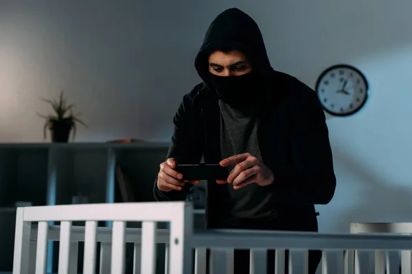 Criminal in mask standing near crib and taking photo — Stock Photo