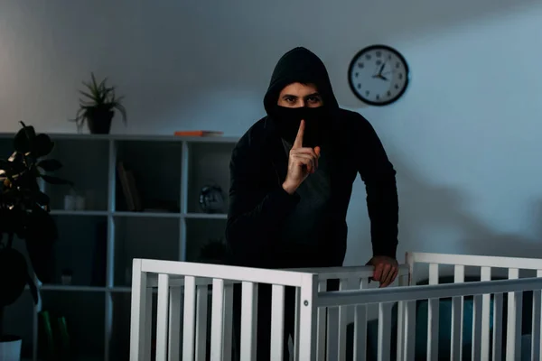 Kidnapper in mask and black hoodie showing hush sign beside crib — Stock Photo