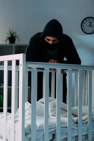 Pensive kidnapper standing in dark room and looking in crib — Stock Photo