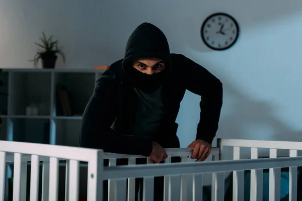 Angry kidnapper in black mask standing near crib and looking at camera — Stock Photo