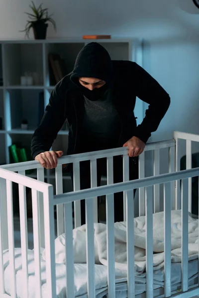 Criminel masqué debout près de la crèche et regardant bébé — Photo de stock