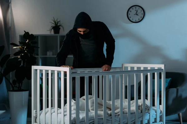 Kidnapper in black clothes standing in dark room and looking in crib — Stock Photo